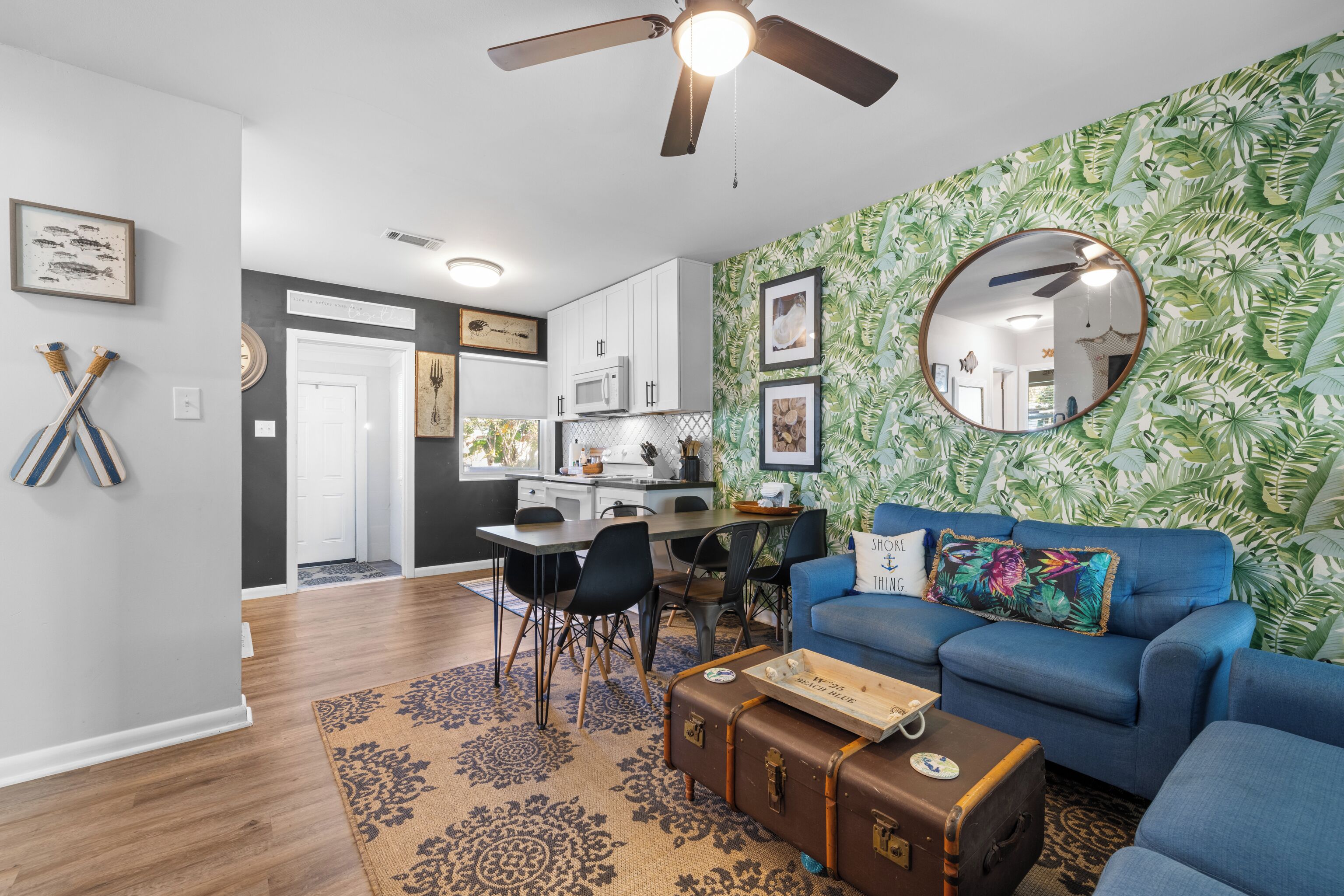 A modern living room with a blue sofa, a dining area with a kitchen in the background, and a tropical leaf-patterned wallpaper.