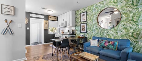 A modern living room with a blue sofa, a dining area with a kitchen in the background, and a tropical leaf-patterned wallpaper.