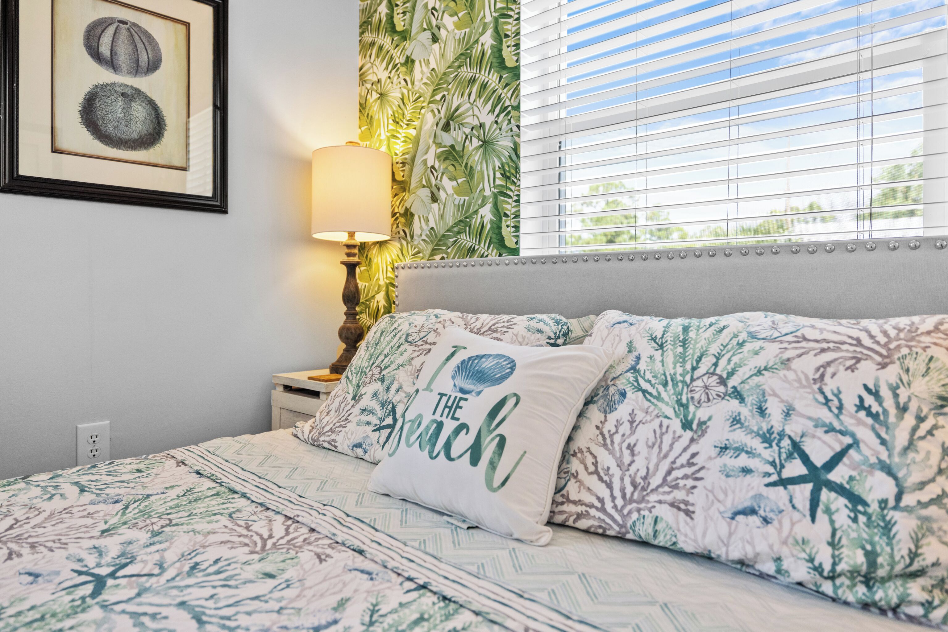 A bedroom with a bed, a nightstand, a lamp, a framed picture, and a window with blinds.