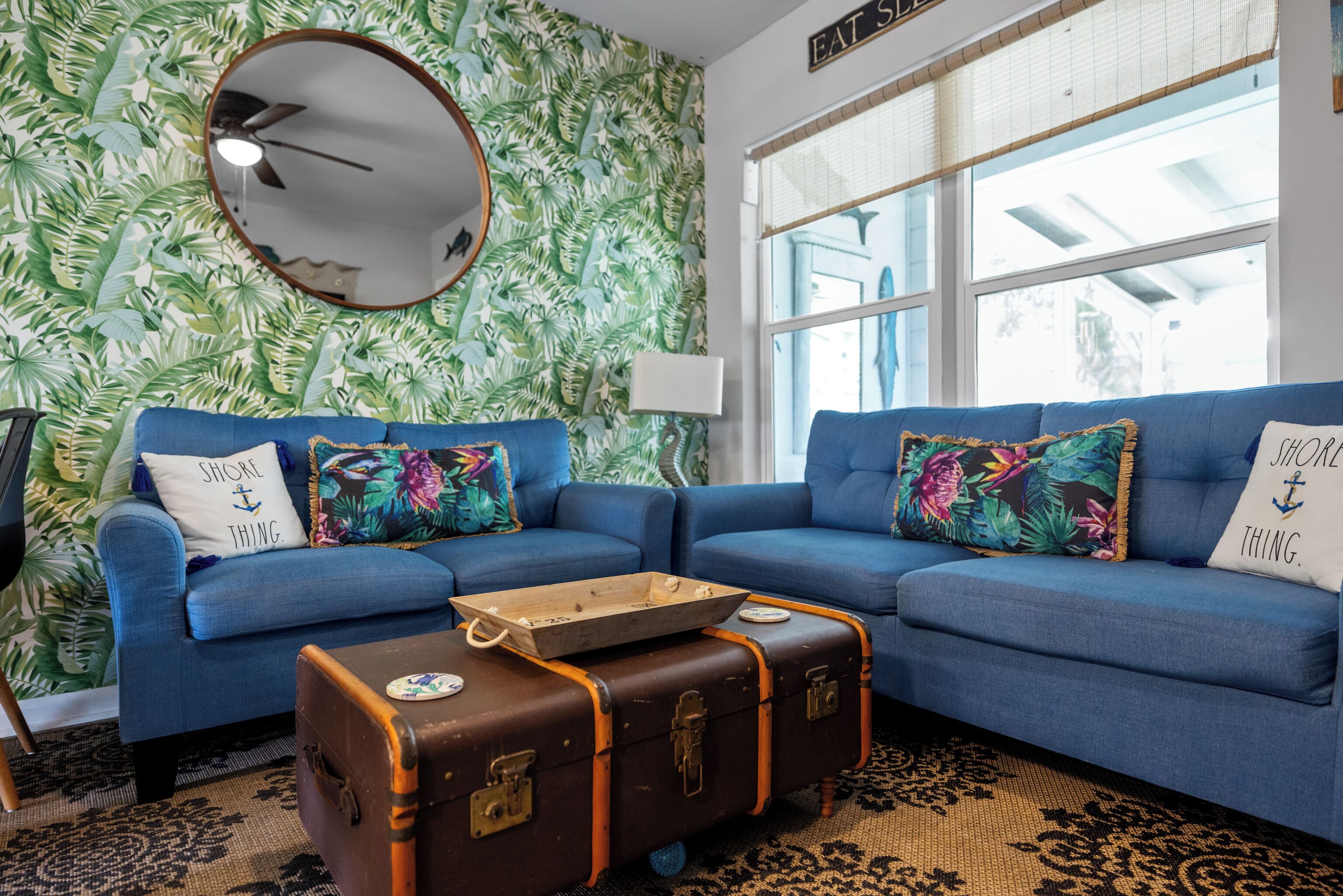 Ein Wohnzimmer mit blauem Sofa, tropischem Wandpapier, Deckenventilator und einem Fenster mit Jalousien.