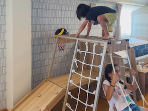 A child climbing a wooden ladder with a netted structure, a basketball hoop, and a bed with a striped bedspread in a room with a patterned wall. - THE AOCA TOKYO JIYUGAOKA (Setagaya-ku)