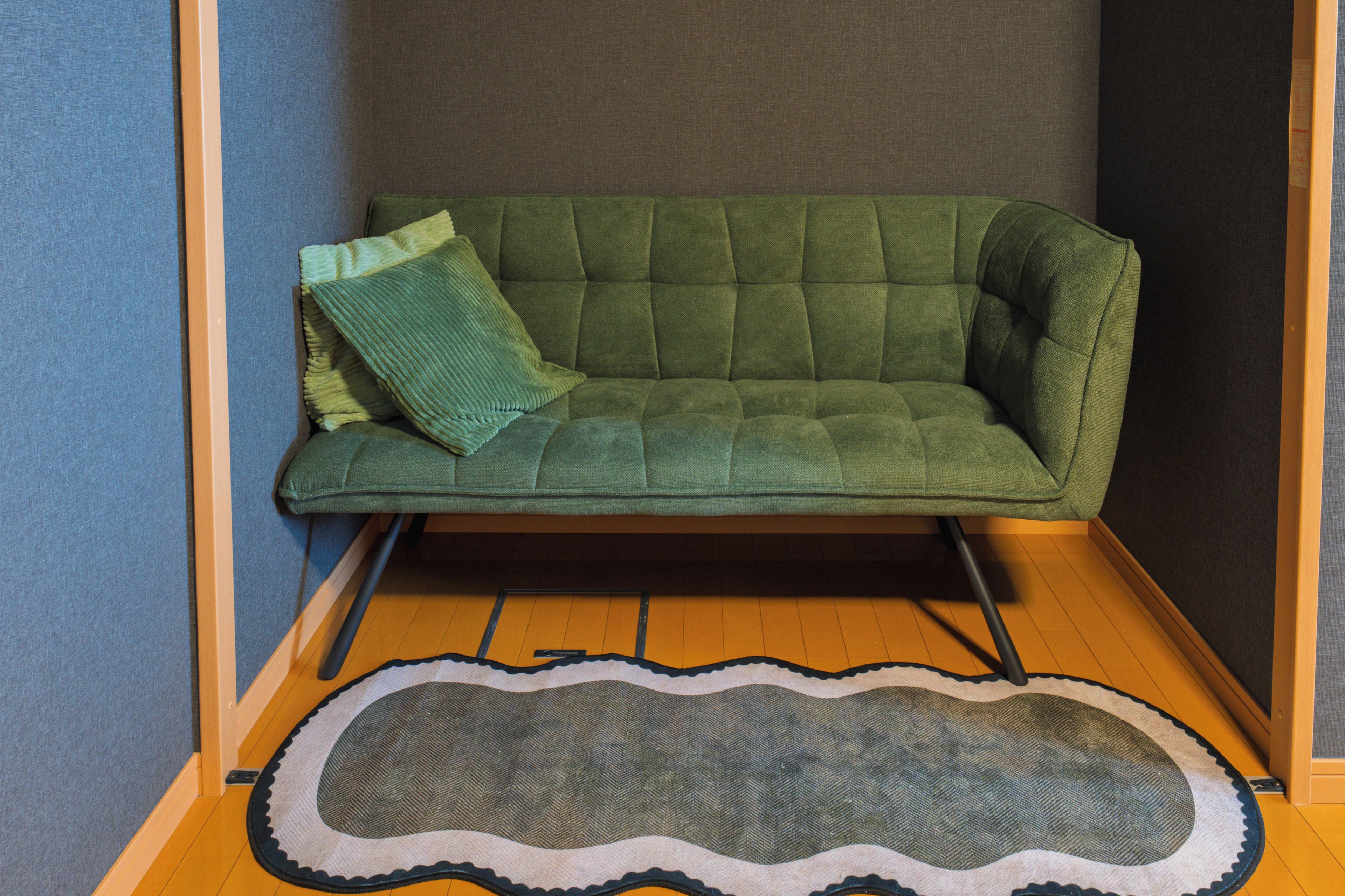 A green sofa with a cushion in a corner of a room with wooden flooring and a patterned rug.