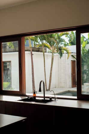A kitchen with a window overlooking a white wall and a small garden with palm trees. - Villa Lurline (Kuta Selatan)