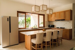 A modern kitchen with wooden cabinets, a marble countertop, and a central island with chairs. - Villa Lurline (Kuta Selatan)