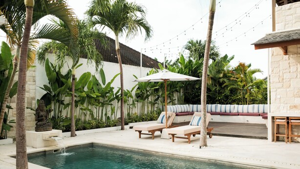 A pool area with lounge chairs, a small waterfall feature, and a stone wall with tropical plants. - Villa Lurline (Kuta Selatan)