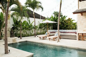 A pool area with lounge chairs, a small waterfall feature, and a stone wall with tropical plants. - Villa Lurline (Kuta Selatan)