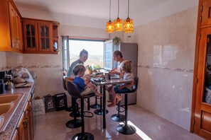 A family dining at a kitchen counter with stools, a refrigerator, and a window with a view. - Detached Beach House near Santa Cruz | Surf, Yoga & Ocean Views | Sleeps 8–10⁠ (Silveira)