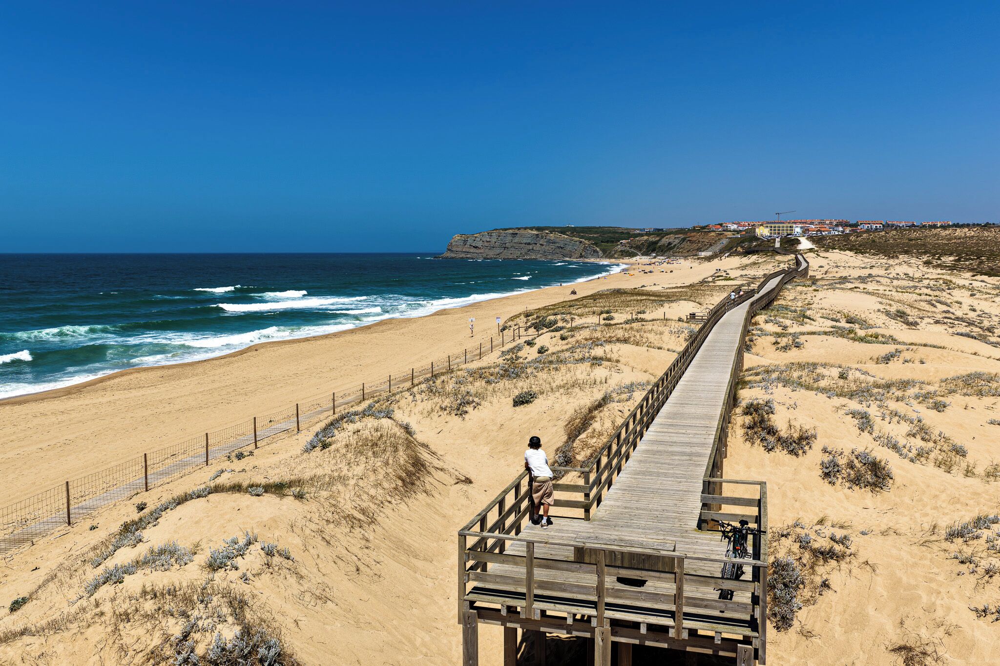 A wooden boardwalk leading to a beach with a person standing at the end.