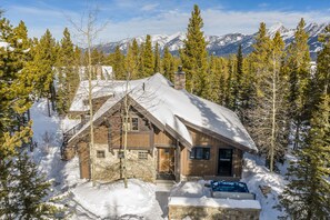 A cabin with a stone facade, wooden exterior, and a snow-covered roof, surrounded by evergreen trees and mountains in the background. - New Listing! Slopeside at Cowboy Heaven (Big Sky)