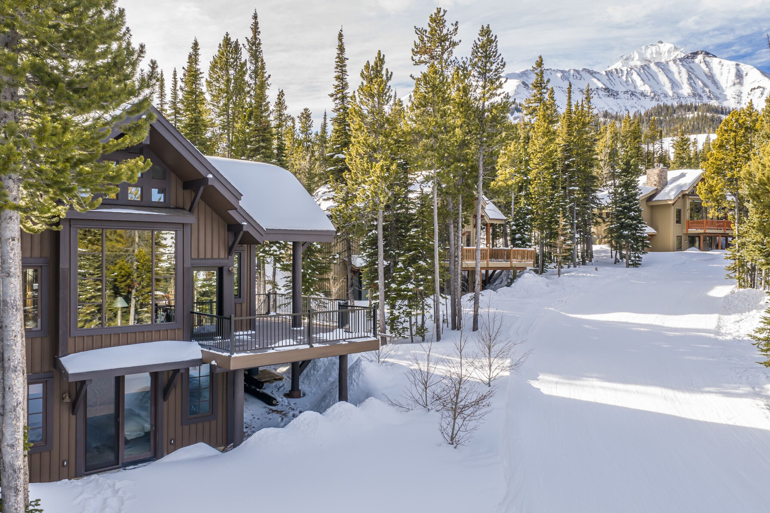 A modern cabin with a deck, surrounded by snow-covered ground and trees, with a mountain in the background.