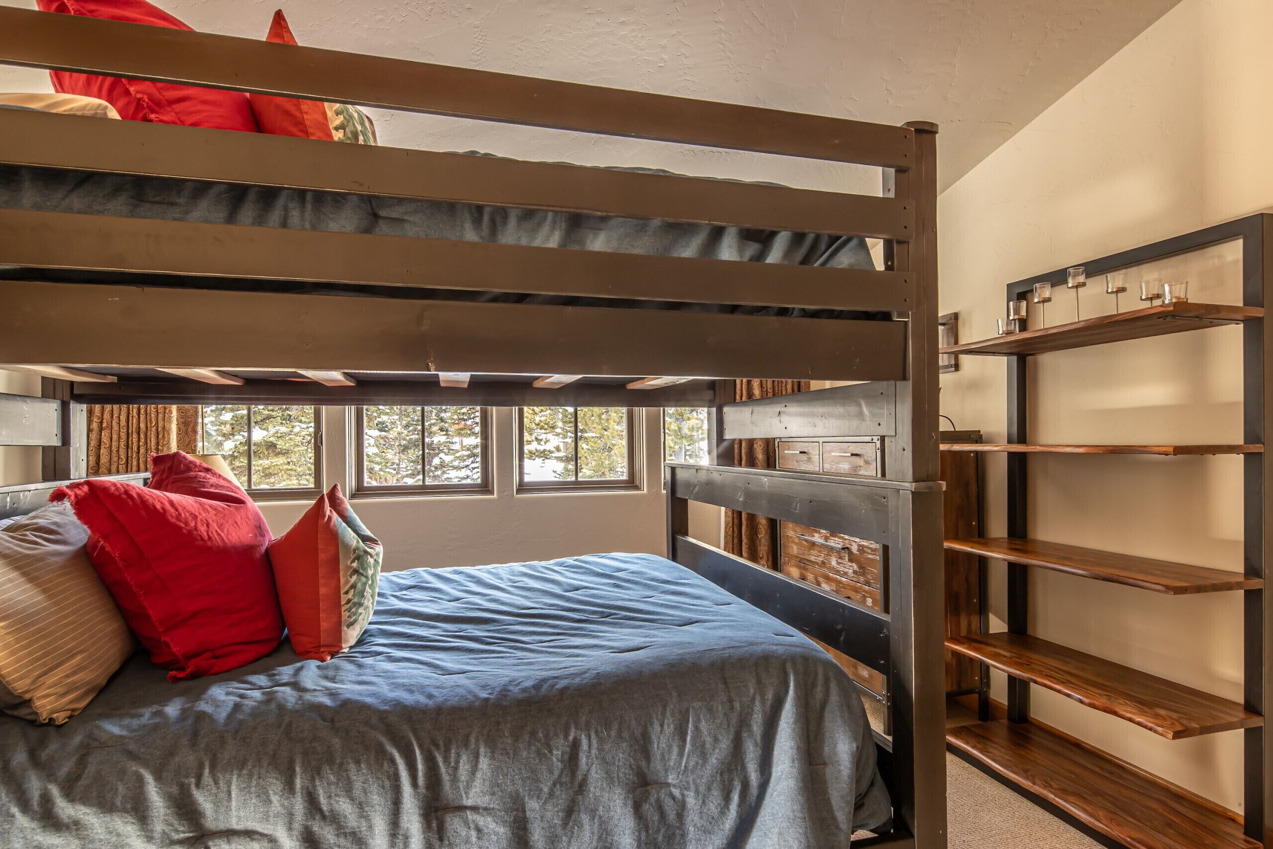 Ein Etagenbett mit blauer Matratze und roten Kissen, eine Holzregaleinheit mit Gläsern sowie ein Fenster mit Blick auf Bäume.