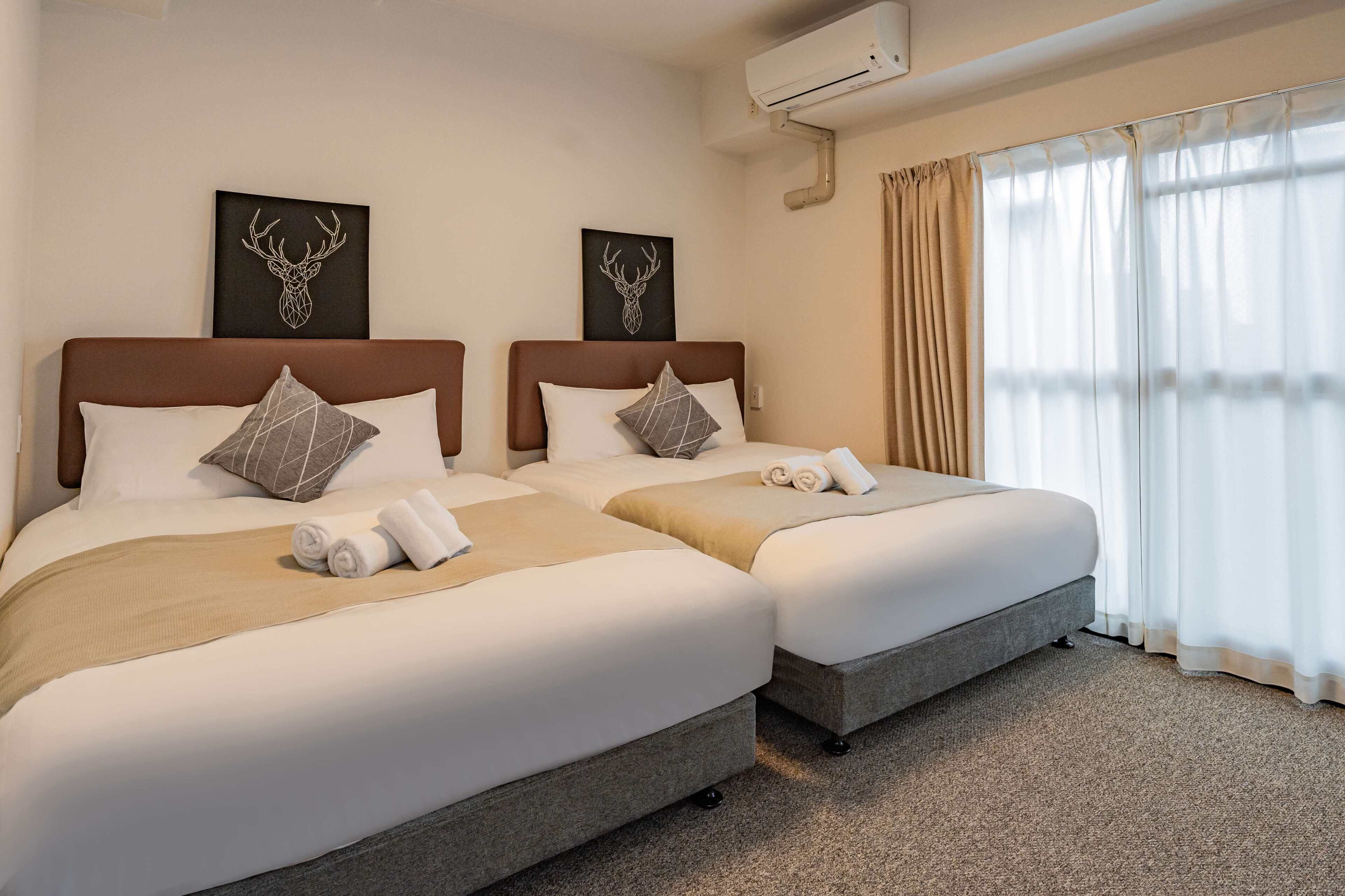 A hotel room with two beds, each with a brown headboard, white bedding, and decorative pillows. There are wall-mounted air conditioning units and a large window with sheer curtains.