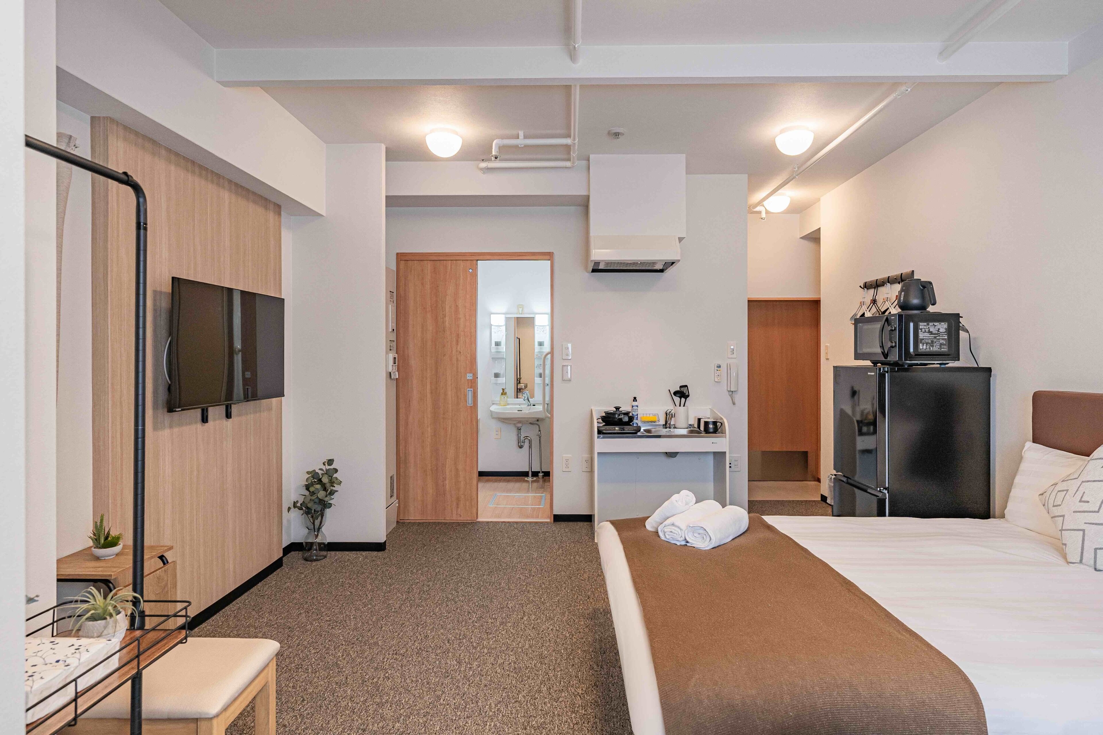 A hotel room with a bed, a desk with a computer, a TV, a refrigerator, and a bathroom visible through an open door.