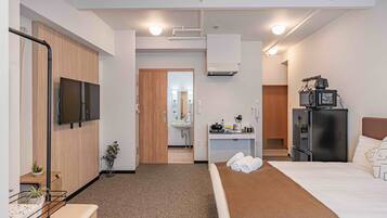 A hotel room with a bed, a desk with a computer, a TV, a refrigerator, and a bathroom visible through an open door.