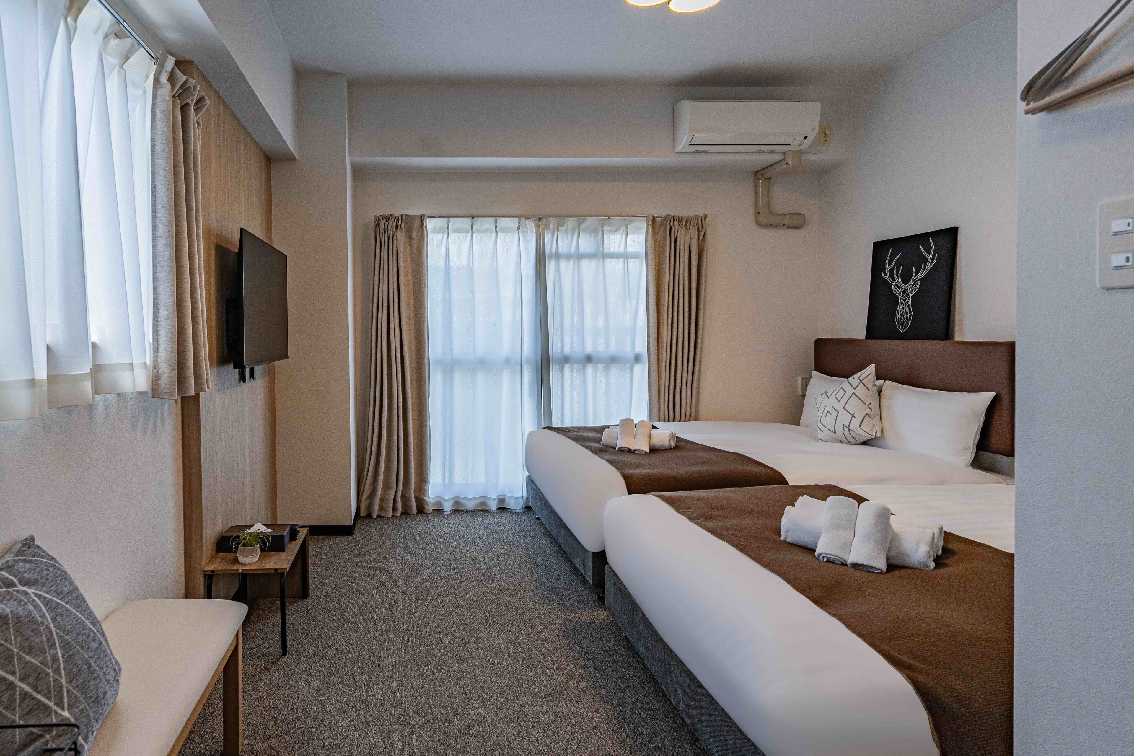 A hotel room with a bed, a TV, a small table with a plant, and a window with curtains.
