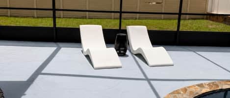Zona junto a la piscina con dos sillones de descanso y una mesita, rodeada por un porche cubierto con vistas a un jardín y una piscina.