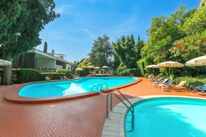 Ein Schwimmbecken mit Liegestühlen und Sonnenschirmen, umgeben von Bäumen und einem Pflasterweg.