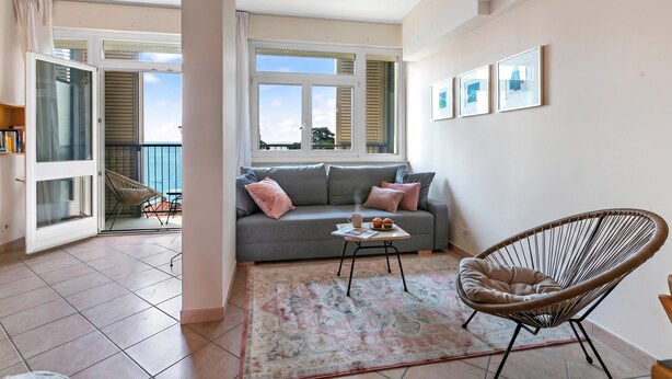 Ein Wohnzimmer mit grauem Sofa, einem Korbstuhl, einem Couchtisch und Meerblick durch große Fenster.