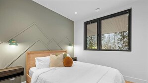 Une chambre à coucher avec un lit, une table de chevet, une lampe et une fenêtre donnant sur des arbres.