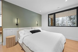 A modern bedroom with a large bed, two wall-mounted lights, a window with a view of trees, and a wooden floor. - Luxury Riverside Retreat near Whistler (Whistler)