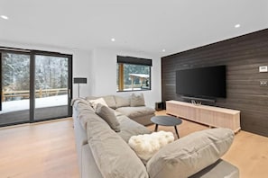 A modern living room with a large flat-screen TV, a sectional sofa, and a wooden entertainment unit. - Luxury Riverside Retreat near Whistler (Whistler)