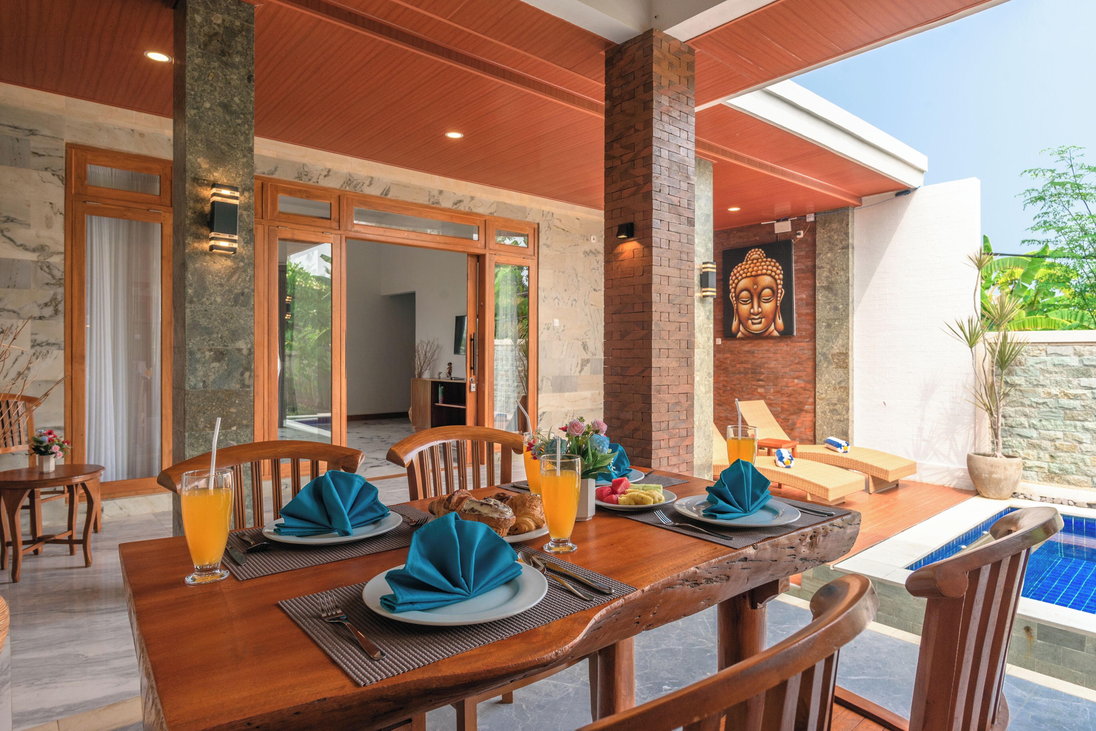 Un espace repas avec vue sur la piscine, comprenant une table dressée avec des assiettes, des serviettes et des boissons, ainsi qu’une statue de Bouddha accrochée au mur.