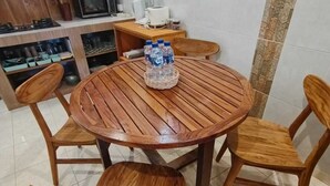 Una mesa redonda de madera con seis sillas, una cesta tejida con botellas de agua, y una cocina al fondo con armarios y electrodomésticos.