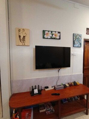 Una sala de estar con un televisor de pantalla plana montado en la pared, una mesa de centro de madera con revistas y un estante pequeño con adornos.