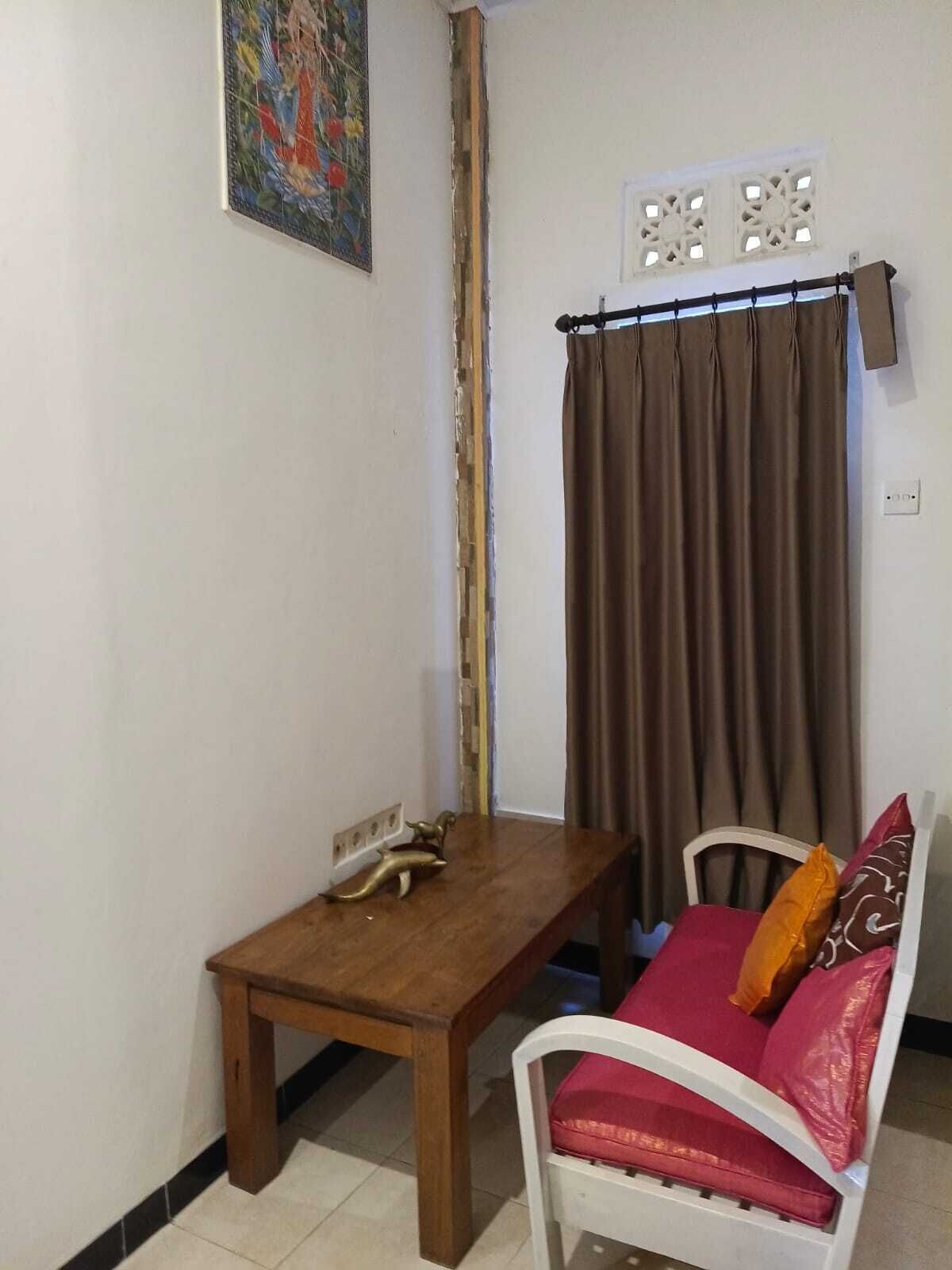 A small room with a wooden table, a white chair with red cushions, a brown curtain, and a decorative wall piece.