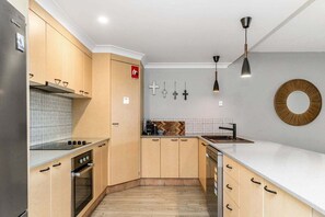 A modern kitchen with wooden cabinets, a built-in oven, a white countertop, and a hanging pendant light. - Indiah - Unit 3, Byron Quarter Apartments (Byron)