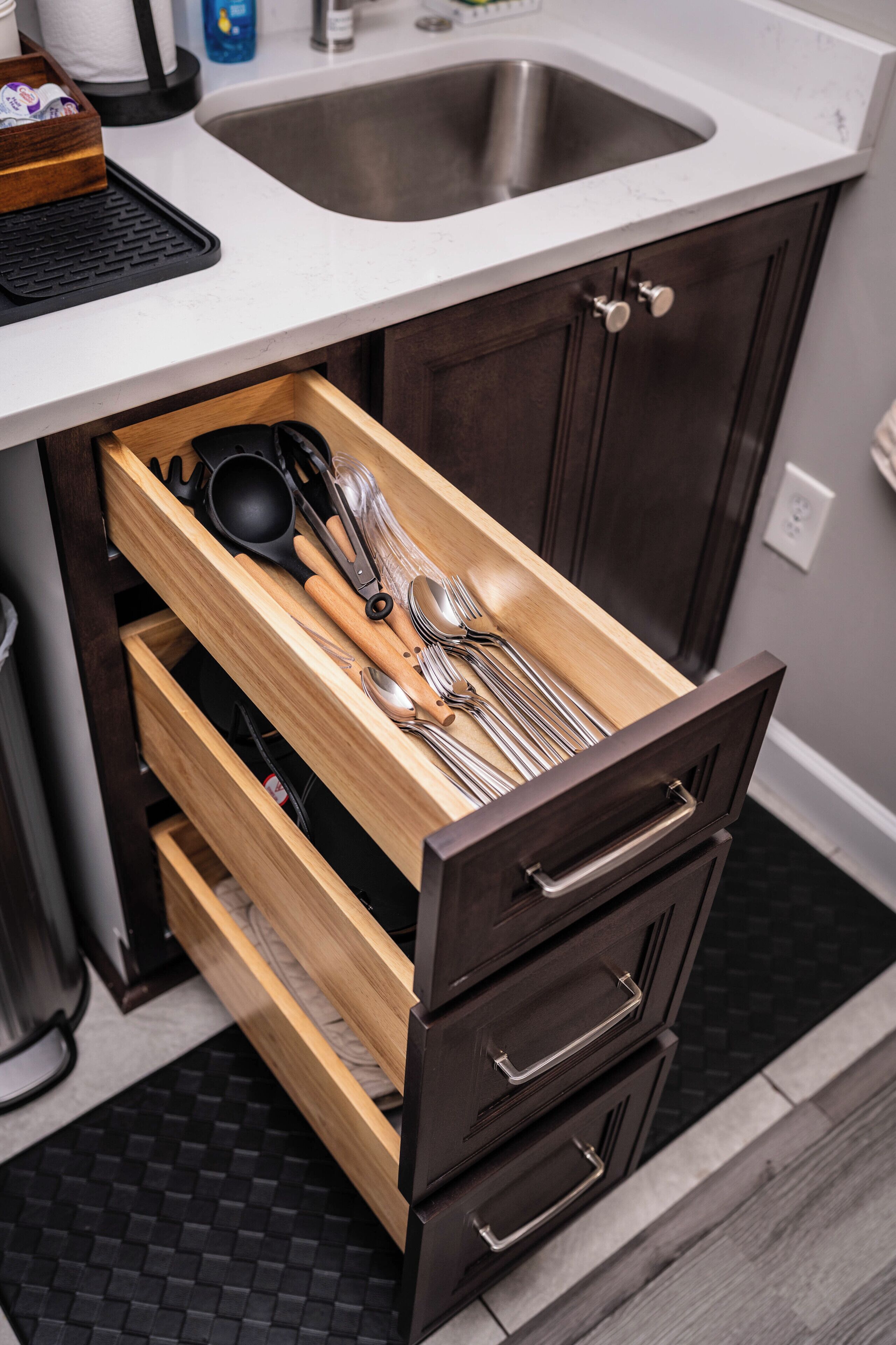 A kitchen with a stainless steel sink, wooden drawer with utensils, and a black tiled backsplash.