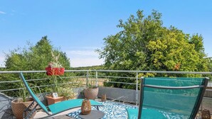 Ein Balkon mit Metallgeländer, Blumentöpfen, einem Tisch mit Stühlen und Blick auf Bäume und Hügel.