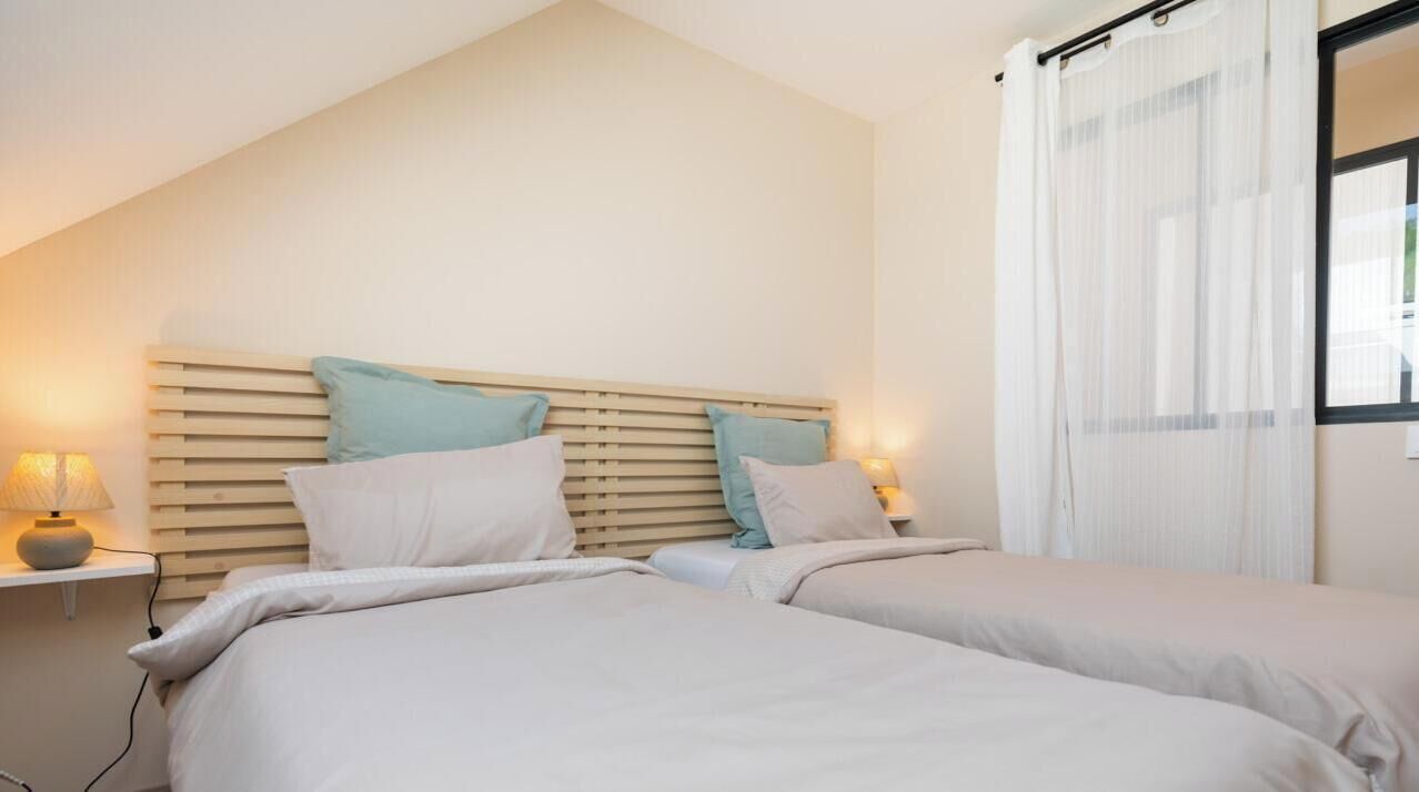 A bedroom with two beds, each with a headboard, bedside lamps, and a window with curtains.