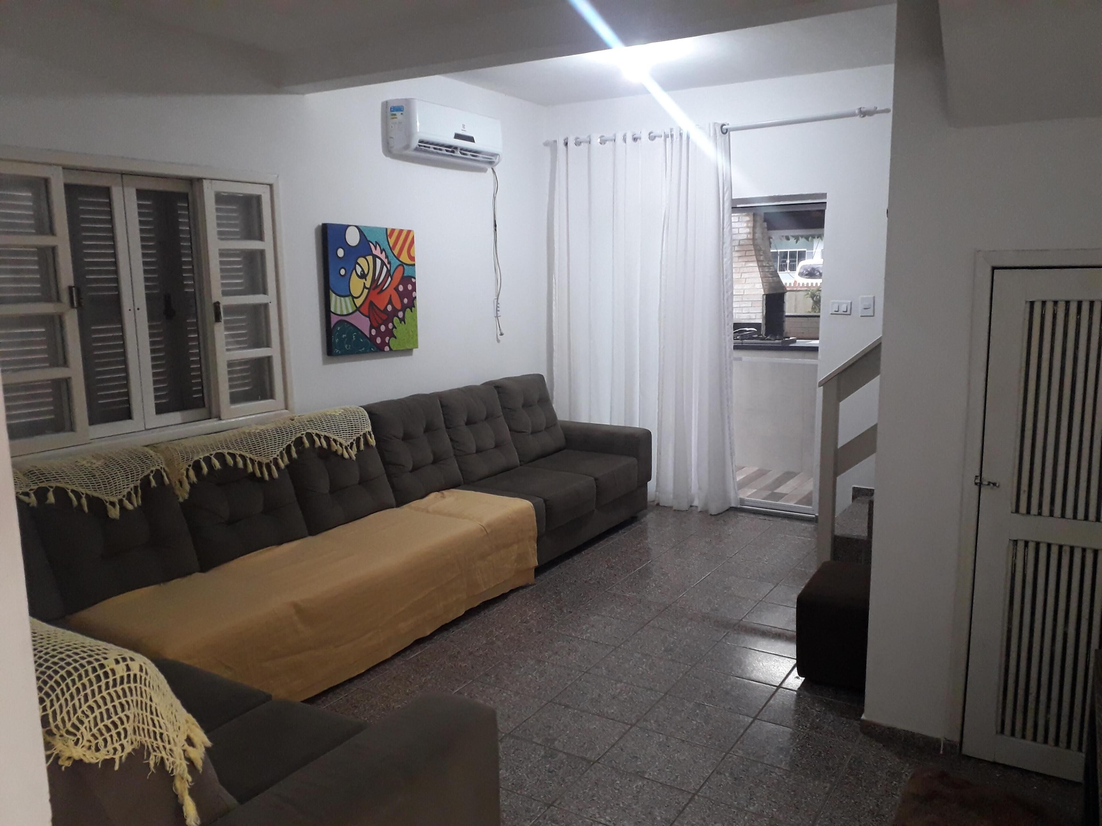 A living room with a sectional sofa, a painting on the wall, an air conditioning unit, and a sliding glass door leading to a balcony.