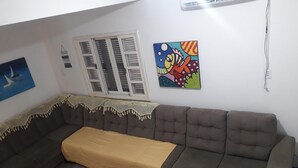 Een woonkamer met een bruine bank, een gele plaid, een raam met luiken, en een kleurrijk schilderij aan de muur.