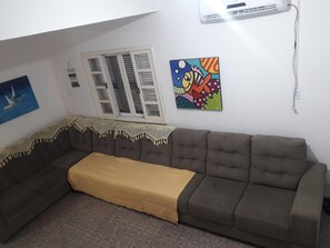 A living room with a brown sofa, a yellow throw blanket, a window with shutters, and a colorful painting on the wall. - Two-story house with pool and small soccer field in Itapoá-SC (Itapoá)