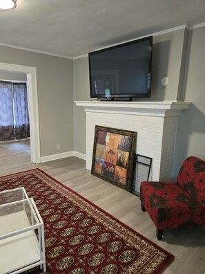 A living room with a flat-screen TV mounted above a white fireplace, a red floral sofa, a patterned rug, and a glass coffee table.