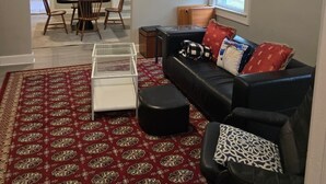 A living room with a black leather sofa, a glass coffee table, and a red patterned rug. There is a window with blinds, a staircase leading to another room, and a dining area visible in the background.