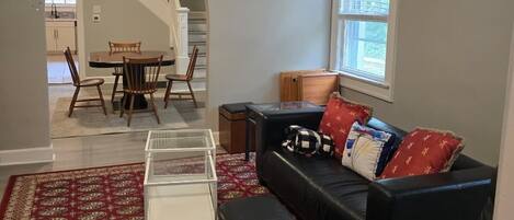 A living room with a black leather sofa, a glass coffee table, and a red patterned rug. There is a window with blinds, a staircase leading to another room, and a dining area visible in the background.