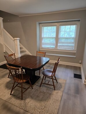 A dining area with a wooden table and chairs, a window with blinds, and a staircase with white railings.