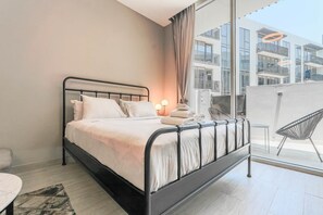A modern bedroom with a metal bed frame, a bedside table with a lamp, and a view of a building through large windows. - HAVN - Studio in Q Gardens Boutique, Arjan (Dubai)