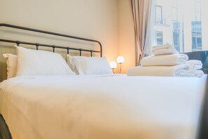 A neatly made bed with white linens and pillows, a metal headboard, and a stack of folded towels on the side. - HAVN - Studio in Q Gardens Boutique, Arjan (Dubai)