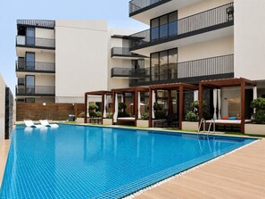 A swimming pool with lounge chairs, a wooden deck, and a building with balconies. - HAVN - Studio in Q Gardens Boutique, Arjan (Dubai)