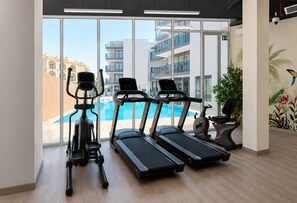 A gym with treadmills, an elliptical, and a stationary bike, with a view of buildings and a pool outside. - HAVN - Studio in Q Gardens Boutique, Arjan (Dubai)