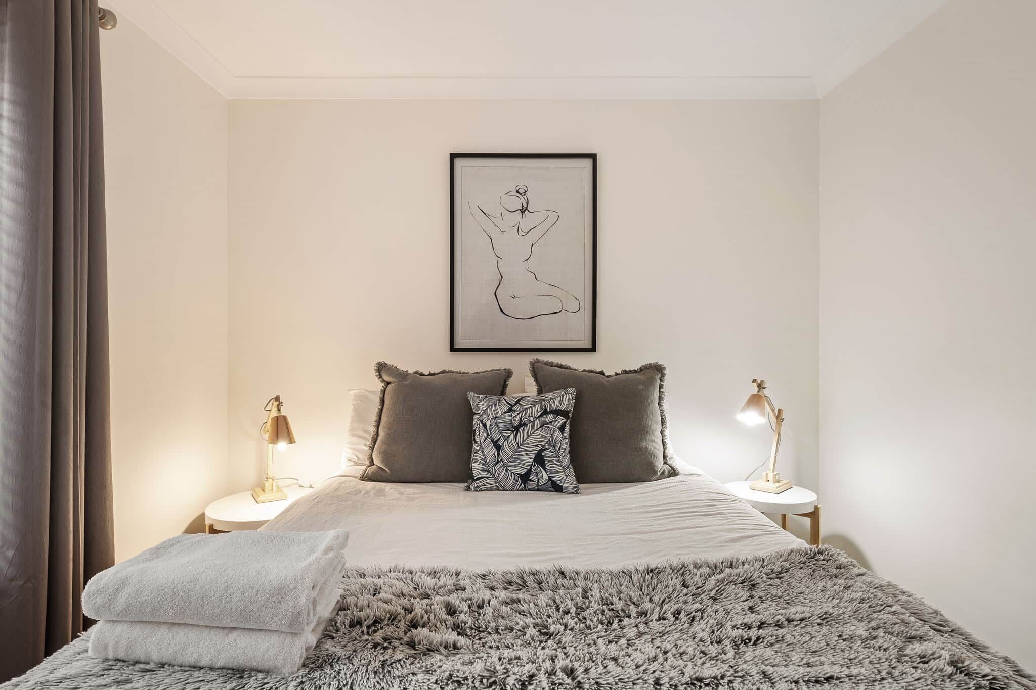 A bedroom with a bed, two bedside lamps, a framed artwork, and a fluffy rug.