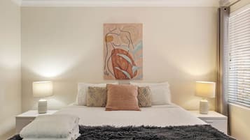 A bedroom with a bed, two bedside tables, a large abstract painting, and a window with blinds.