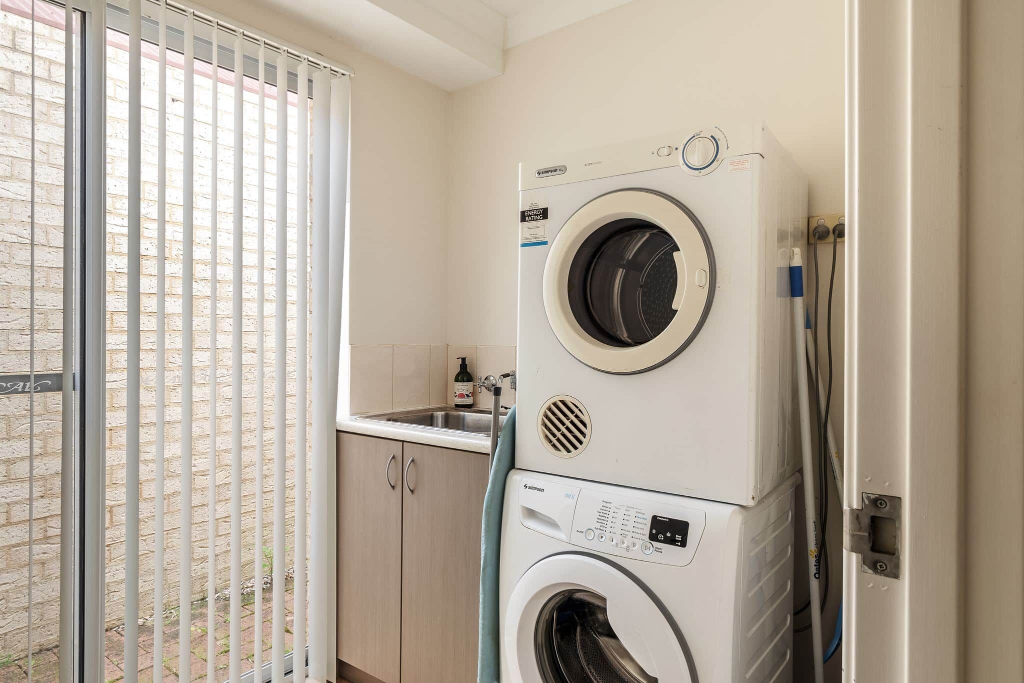 Une buanderie équipée d’un lave-linge et d’un sèche-linge superposés, d’un évier et d’une fenêtre avec des stores.
