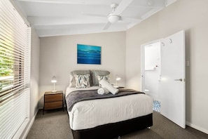 Ein Schlafzimmer mit Bett, Nachttischen, Deckenventilator, Tür und Fenster mit Jalousien.