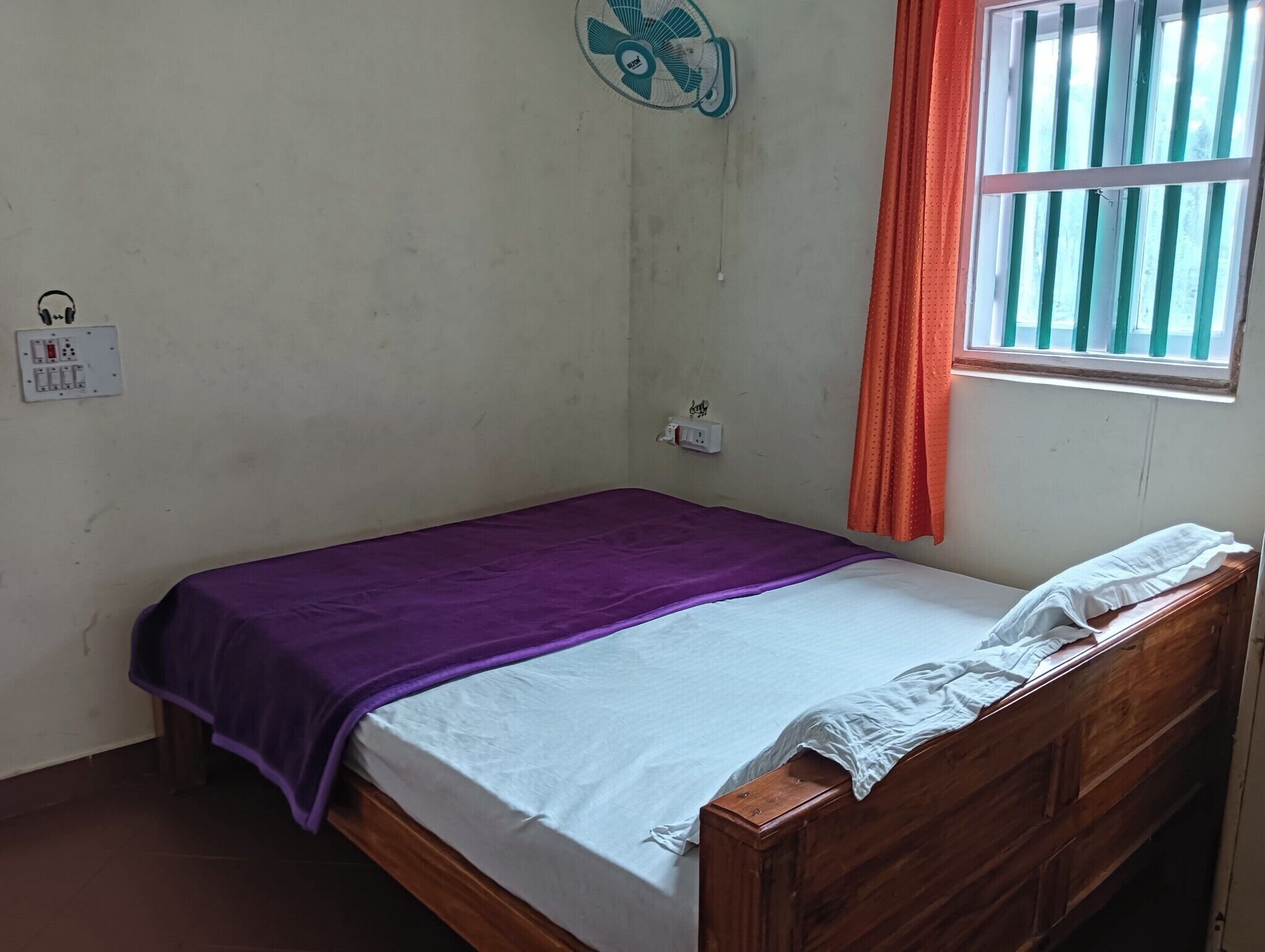Una cama individual con una manta morada, una sábana blanca y un cabecero de madera. Hay un enchufe y una toma de corriente en la pared.