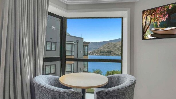 A room with a large window offering a view of a lake and mountains, a round table with two chairs, and a painting on the wall. - Lakeside Luxury for Two | Modern Oak Shore Retreat (Queenstown)